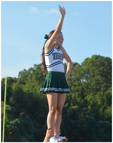 A person wearing a cheerleading uniform with a green skirt and white top is being lifted by another person outdoors with trees and a clear sky in the background. Description generated by AI