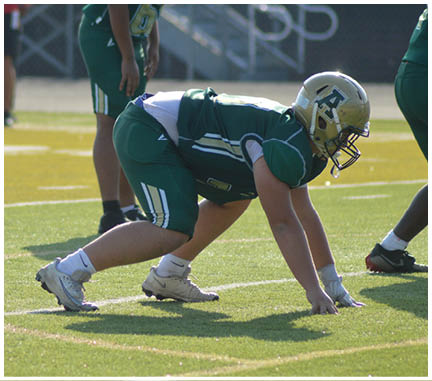 A person in green sports gear is crouched in a ready position on a football field with other people standing in the background. Description generated by AI