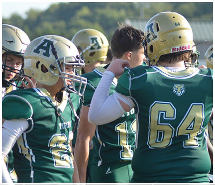 A group of people wearing green and gold football uniforms and helmets are gathered closely together outdoors. Description generated by AI
