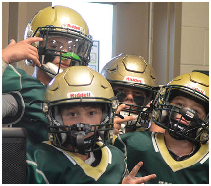Two people wearing green football uniforms and gold helmets are indoors, with one person making a hand gesture near their face. Description generated by AI