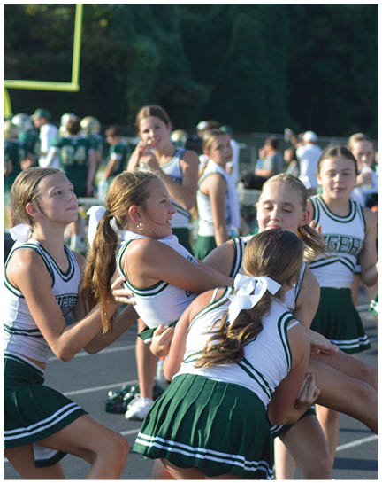 A group of people in green and white cheerleading uniforms are outdoors, with one person being lifted by others while the rest stand nearby on a paved surface. Description generated by AI