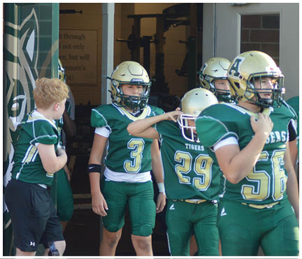 Children wearing green and gold football uniforms and helmets are standing and walking near a brick building entrance. Description generated by AI