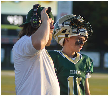 A person wearing a headset stands next to a person in a football uniform with a helmet resting on their head, both looking intently in the same direction outdoors. Description generated by AI