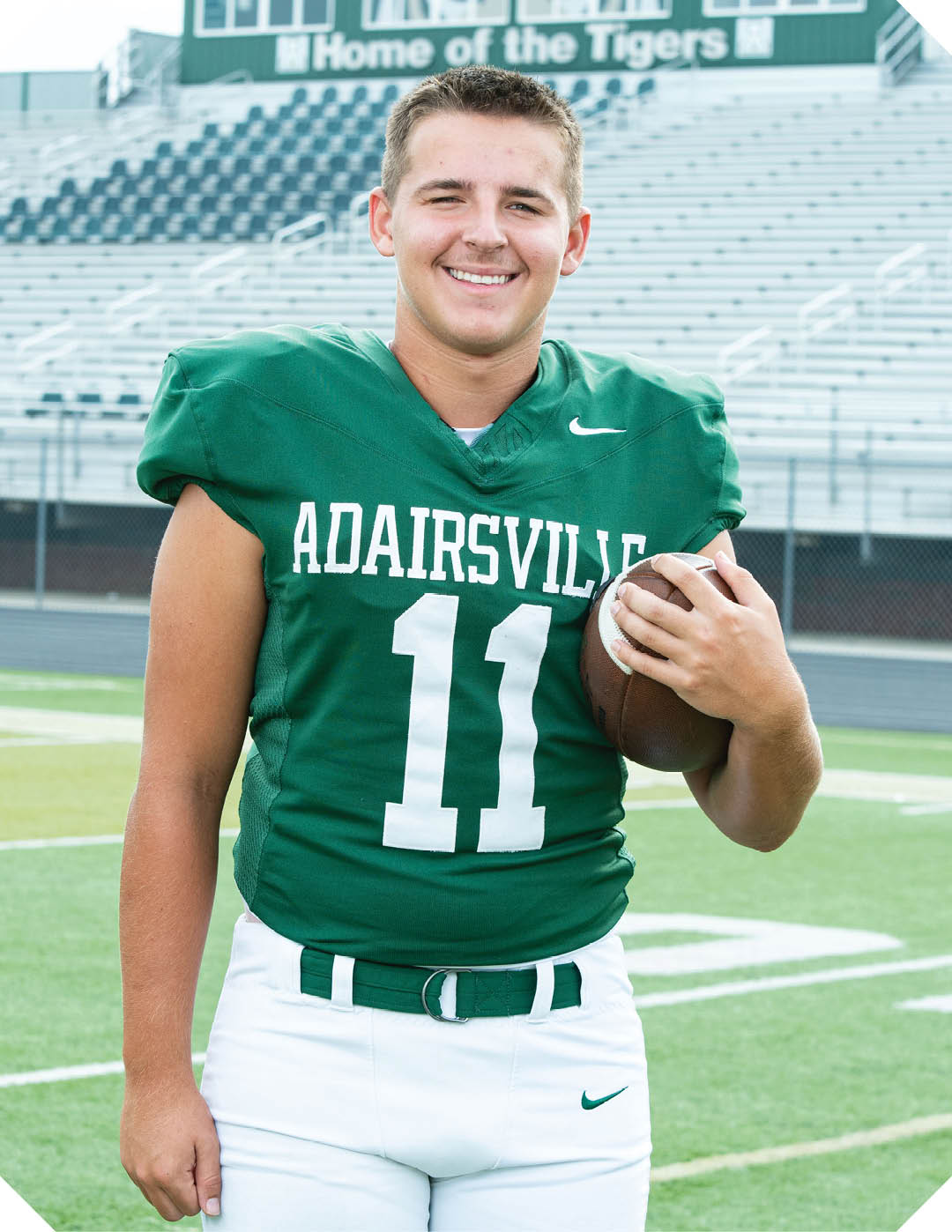 A person wearing a green football jersey with the number 11 and the word Adairsville is standing on a football field holding a football and smiling. Description generated by AI