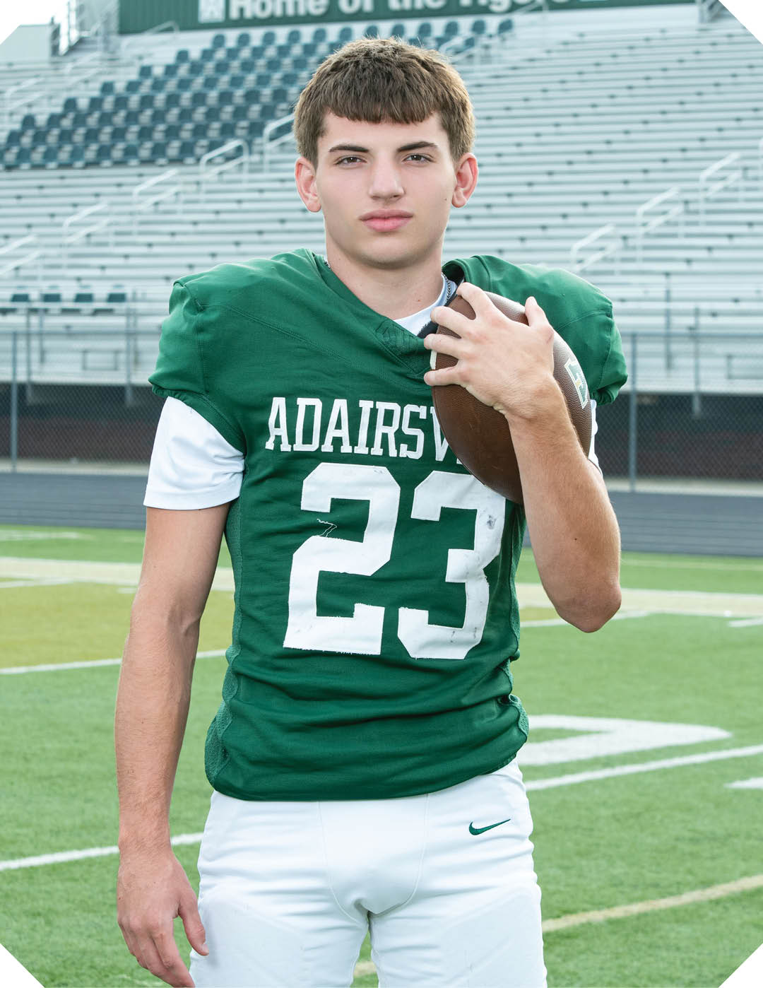 A person wearing a green football jersey with the number 23 and the word Adairsville is standing on a football field holding a football with empty bleachers and a scoreboard that reads Home of the Tigers in the background. Description generated by AI