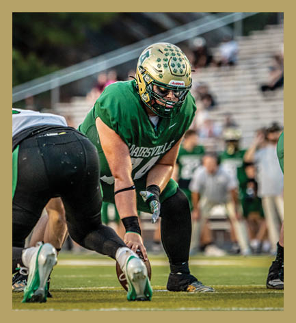 A person wearing a green football uniform and a gold helmet with paw prints is leaning forward on a football field with blurred spectators in the background. Description generated by AI