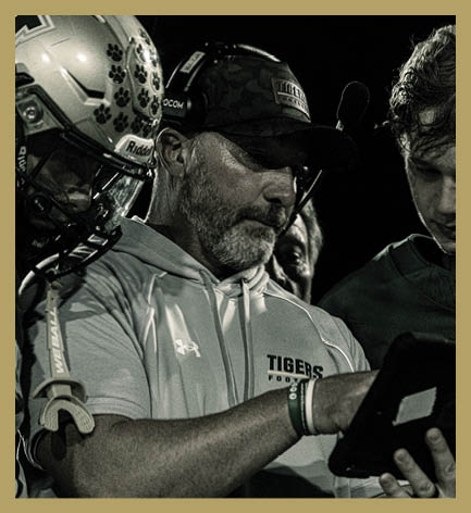 An adult wearing a headset and a cap is showing a tablet to two other people, one wearing a football helmet and the other in a sports jersey, likely during a football game. Description generated by AI