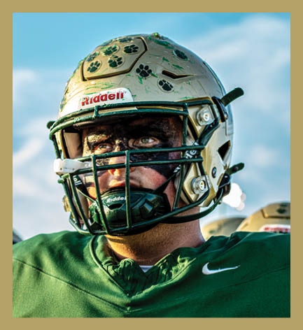 A person wearing a football helmet with paw print stickers and black face paint is dressed in a green sports jersey with a white Nike logo. Description generated by AI
