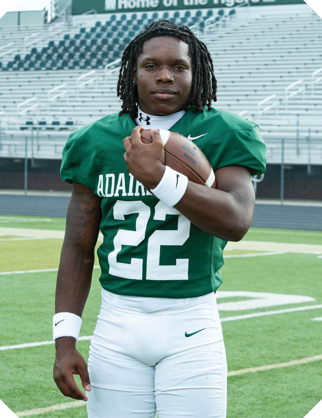 A person wearing a green football jersey with the number 22 and white pants is standing on a football field holding a football close to their chest. Description generated by AI