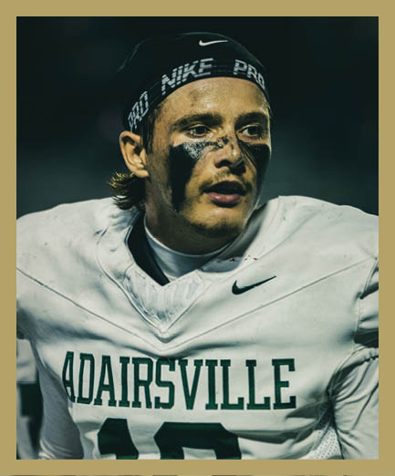 Person wearing a white football jersey with “ADAIRSVILLE" and the number 10 on it, a black Nike headband, and black face paint under the eyes. Description generated by AI