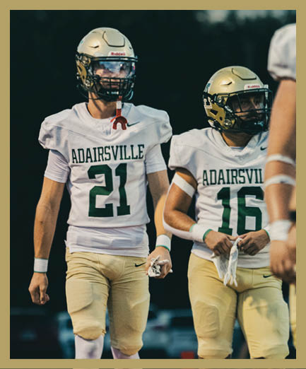 Two people wearing football uniforms with helmets and jerseys numbered 21 and 16 are standing on a field during a game or practice. Description generated by AI