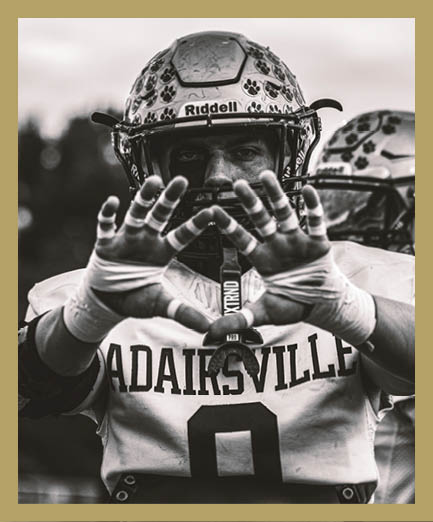 An adult wearing a football helmet and jersey with the word “ADAIRSVILLE" and the number nine is holding their hands up close to the camera in a black and white photo. Description generated by AI