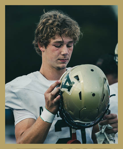 A person wearing a white sports jersey is holding and looking down at a gold football helmet with a green “V" logo. Description generated by AI