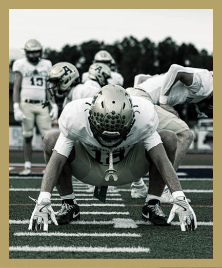A person wearing a white sports jersey is holding and looking down at a gold football helmet with a green “V" logo. Description generated by AI