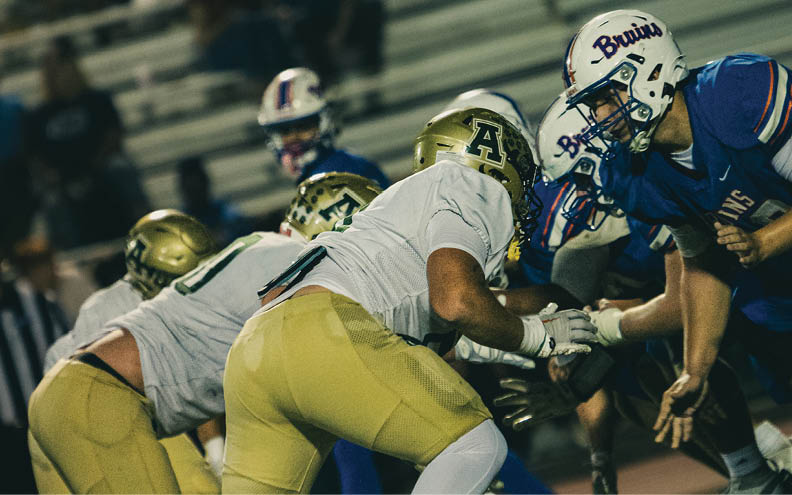 Several people in football uniforms are engaged in a play on a field, with some wearing gold helmets and pants and others in blue jerseys and helmets. Description generated by AI