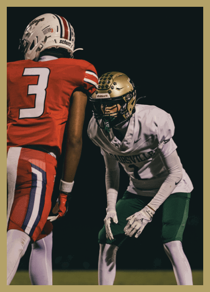 A person wearing a gold football helmet, white jersey, green pants, and white gloves is crouched in a ready position on a dark football field. Description generated by AI