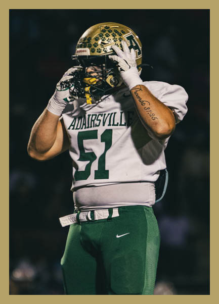 A person wearing a football uniform with the number 51 and a helmet stands on a field adjusting their helmet during a nighttime game. Description generated by AI