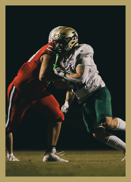 Two people in football uniforms are engaged in a physical play on a dark field, with one wearing red and the other wearing white and green. Description generated by AI