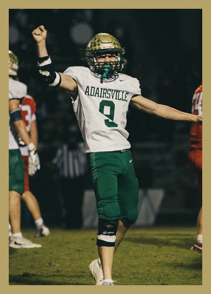 A person wearing a football uniform with the name “ADAIRSVILLE" and number 9 is celebrating on a football field at night with arms raised. Description generated by AI