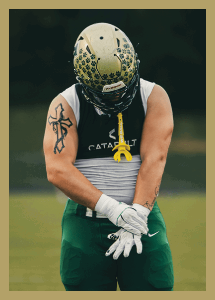 A person wearing a football helmet, gloves, and athletic gear stands on a field with arms crossed and head bowed. Description generated by AI