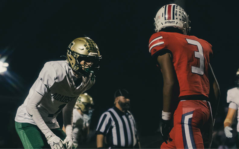 Two people in football uniforms are facing each other on a field at night with a referee standing in the background. Description generated by AI