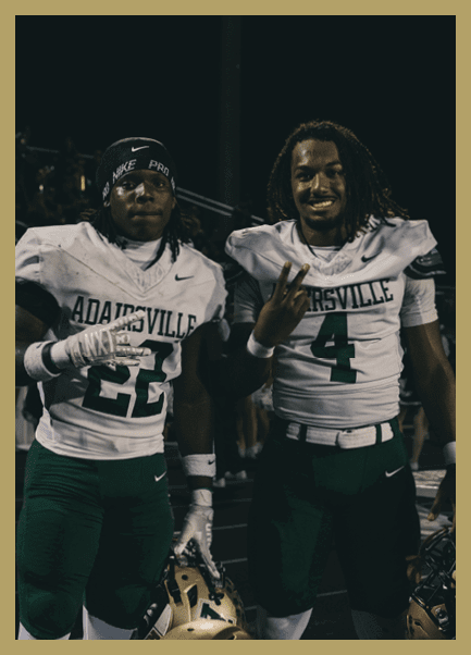 Two adults wearing football uniforms with the name “Anahuacsville" pose together at night, one making a hand gesture and the other smiling. Description generated by AI