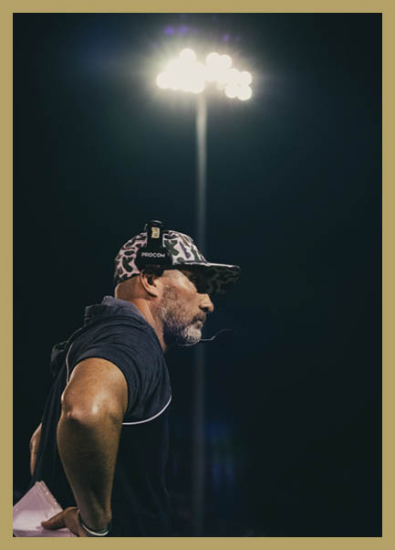 Person wearing a headset and a patterned cap is standing outdoors at night under bright stadium lights. Description generated by AI