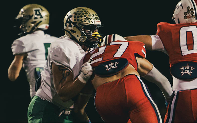 Two people in green and white football uniforms are engaged in a play against two people in red and white football uniforms on a dark field. Description generated by AI