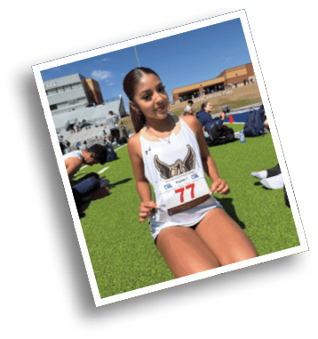 Person wearing a white athletic jersey with a winged logo and holding a race bib numbered 77 at an outdoor track and field event. Description generated by AI