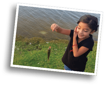 A child wearing a black shirt and denim shorts is smiling and holding up a small fish caught on a fishing line near a grassy shore by the water. Description generated by AI