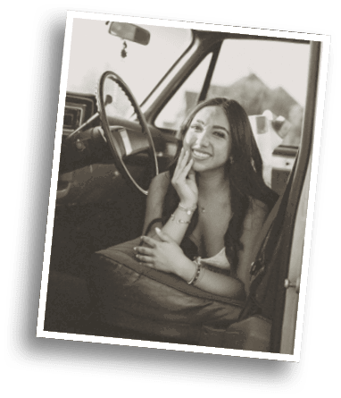 A person with long hair is smiling and resting their face on one hand while sitting inside a vintage car with a steering wheel visible. Description generated by AI