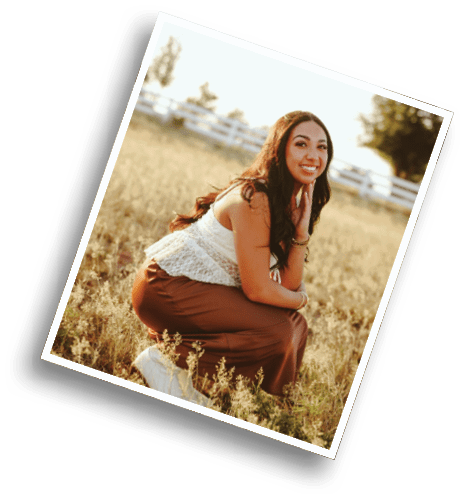 A person with long hair wearing a white sleeveless top and brown pants is sitting in a grassy field smiling with a white fence and trees in the background. Description generated by AI