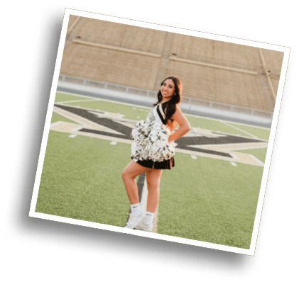 A person wearing a cheerleading uniform and holding pom-poms stands on a football field with empty bleachers in the background. Description generated by AI