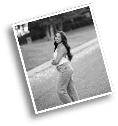 A person with long hair wearing a sleeveless top and jeans is standing outdoors on a path with trees in the background, smiling and looking over their shoulder. Description generated by AI