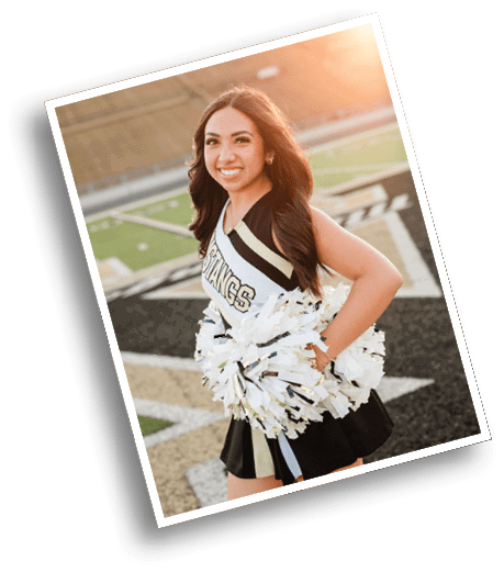 A person with long hair wearing a cheerleading uniform holding white and gold pom-poms stands on a sports field smiling. Description generated by AI