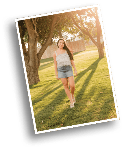 A person with long hair wearing a sleeveless top and denim skirt is walking on grass in a sunlit park with large trees casting long shadows. Description generated by AI