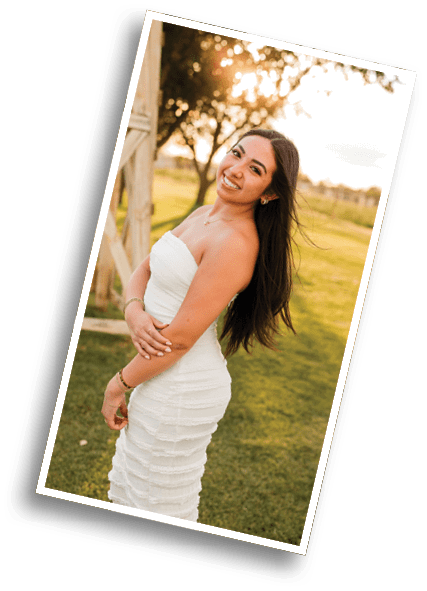 A person with long flowing hair wearing a strapless white dress is smiling outdoors with a blurred natural background. Description generated by AI