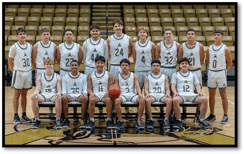 A group of basketball players in white uniforms pose together in a gymnasium with some standing and others sitting on benches, with one player holding a basketball. Description generated by AI