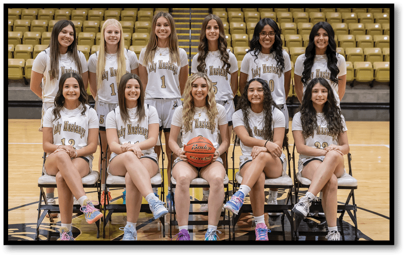 A group of people wearing matching basketball uniforms pose seated and standing on a basketball court with one person holding a basketball in the center. Description generated by AI