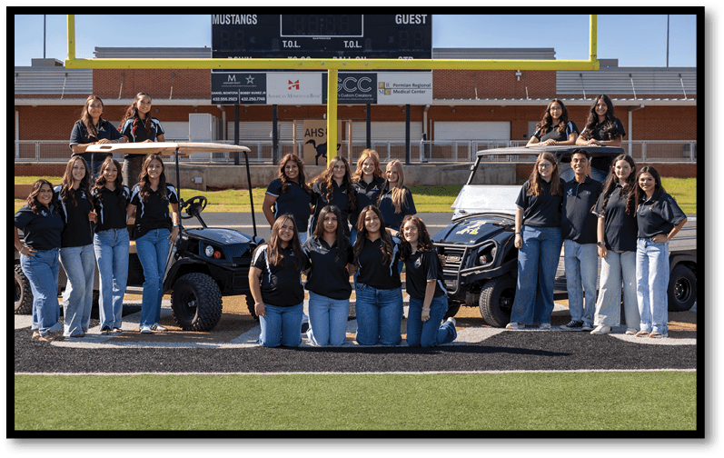 A group of adults wearing matching dark shirts and jeans are posing together on a sports field in front of two utility vehicles and a scoreboard. Description generated by AI