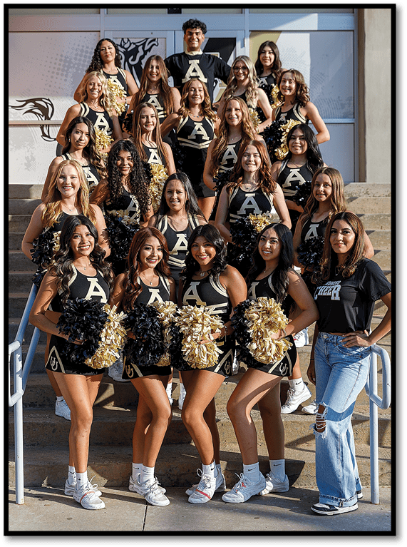 A group of people wearing matching black and gold cheerleading uniforms with pom-poms pose on outdoor steps alongside one person in casual clothing. Description generated by AI