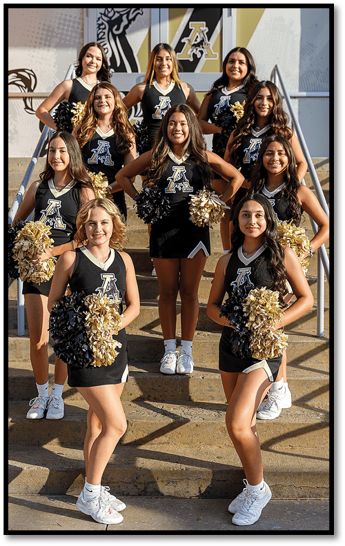 A group of people in matching cheerleading uniforms with pom-poms pose on outdoor steps in front of a building with a large logo. Description generated by AI