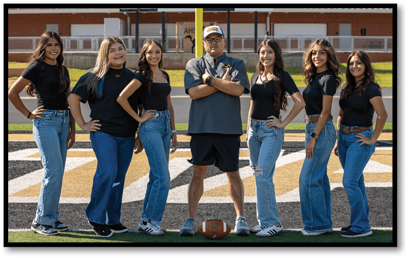 A group of six people wearing casual black tops and jeans stand on a sports field with one person in the center wearing a cap, shorts, and a polo shirt, posing confidently. Description generated by AI