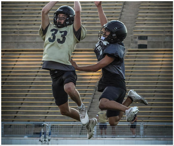 Two people wearing football helmets and athletic gear are jumping in the air on a field with empty bleachers in the background. Description generated by AI