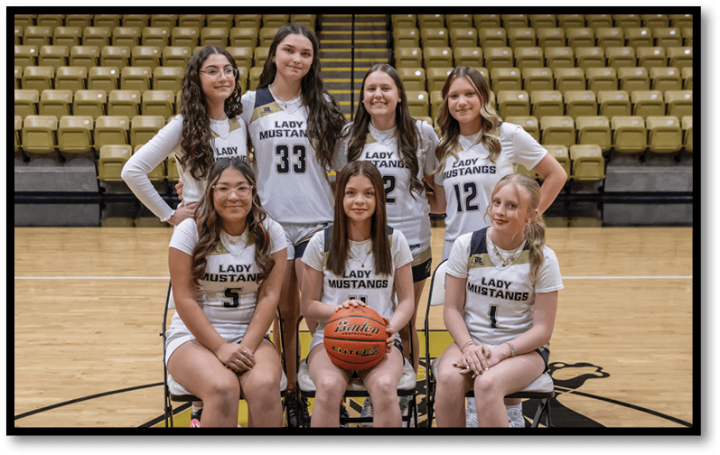 Seven people wearing sports uniforms with “Lady Mustangs" printed on them pose on a basketball court with three seated holding a basketball and four standing behind them in front of empty bleachers. Description generated by AI