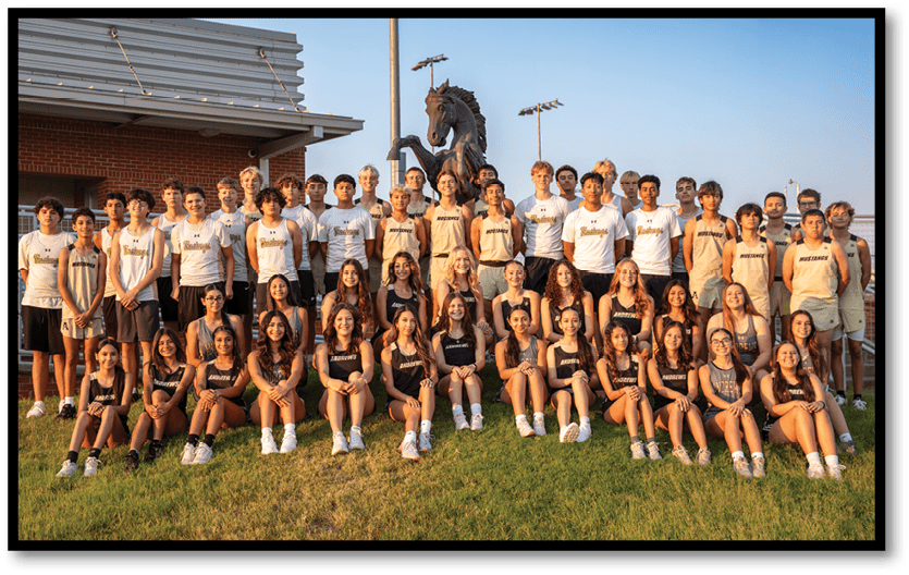 A large group of people wearing matching athletic uniforms is posed outdoors on grass in front of a building and a statue of a horse. Description generated by AI