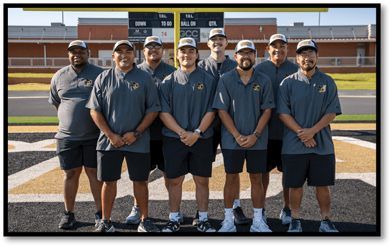 Seven adults wearing matching dark gray polo shirts and shorts stand on a football field in front of a scoreboard and goalpost, posing for a group photo. Description generated by AI