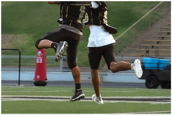 Two people wearing black football uniforms and helmets jump in the air on a football field near the sideline with bleachers and equipment in the background. Description generated by AI