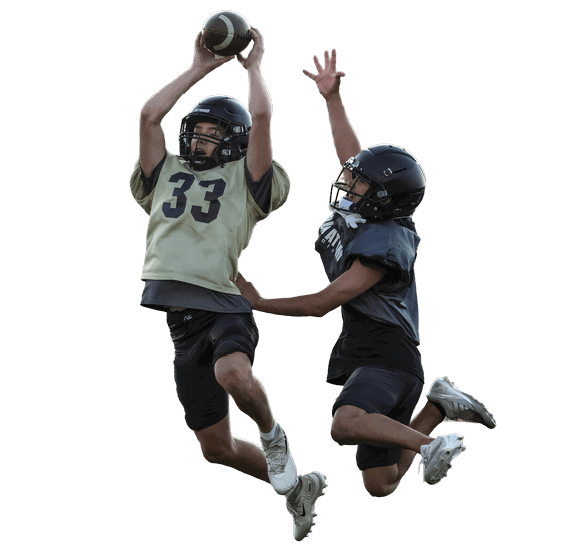 Two people wearing helmets and sports gear are jumping in the air on a football field, with one catching a football while the other reaches up to block or intercept. Description generated by AI