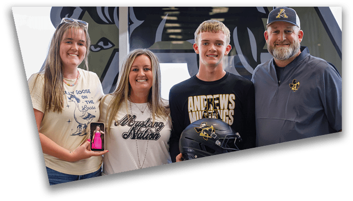 Four people stand closely together smiling, with one holding a phone displaying a small child and another holding a black football helmet with a yellow logo. Description generated by AI